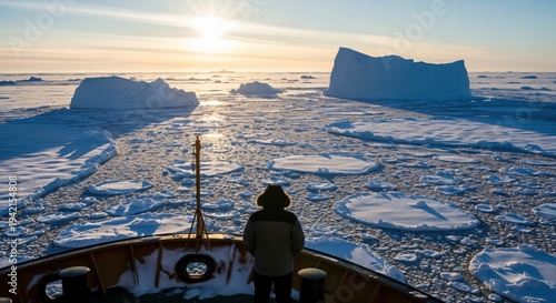 A person in a warm parka standing on the deck of a ship looking at icebergs and pancake ice in the Arctic ocean during a golden sunset