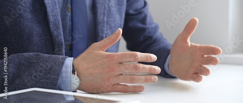 Quadro su tela Businessman wearing blue suit and wristwatch, is sitting at white desk explaining business strategy using hand gestures, with tablet and eyeglasses reflecting on the surface