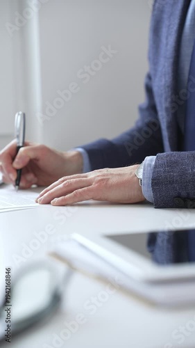 Close up of businessman's hands signing a contract with elegant pen, wearing a blue suit and a wristwatch, sitting at a white desk. Business people concept
