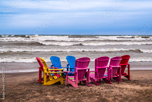 A row of colorful Adirondack chairs arranged in a semicircle on Woodbine Beach in Toronto during a windy spring day with waves.