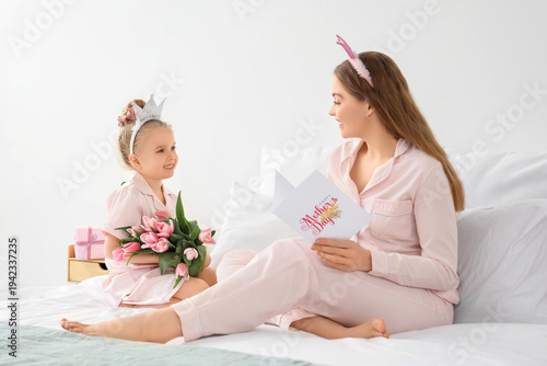 Cute little girl greeting her mom for Mother's Day with tulips and postcard on bed in bedroom