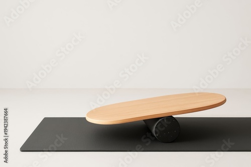 A wooden balance board rests on a black foam roller. It sits on a dark grey mat in a minimalist setting.