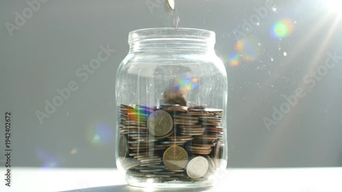 A jar filled with coins on a table with a coin being added.