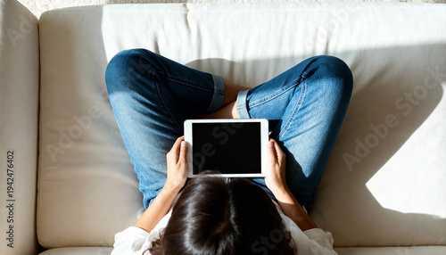 Top view of a woman sitting cross-legged on a sofa holding a tablet. Person relaxing at home with a digital device mockup. Blank black screen for copy space