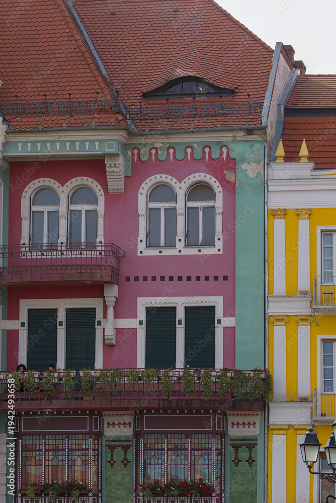 Fototapeta premium Colorful art nouveau house stands on a street in Timisoara Romania. Curved red tiles and arched windows shape the playful facade. Pastel walls and balcony plants soften the street view.