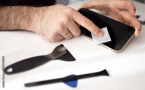 Hands cleaning smartphone display and installing tempered glass screen protector using repair tools. Process of mobile device protection and screen replacement.