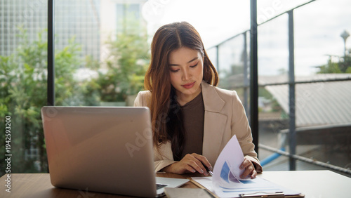 Professional businesswoman reviewing financial charts and documents while working on a laptop in a bright modern.