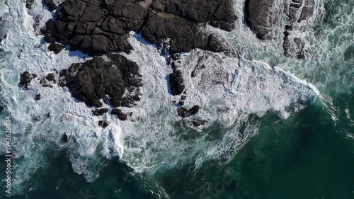 Powerful ocean waves crashing against a rugged rocky shoreline, white foam exploding over uneven stone formations as restless surf meets the coast, captured from above to emphasize texture, force, and