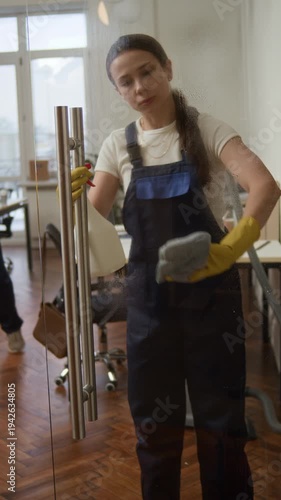 Vertical shot of young Caucasian female cleaner using sprayer and wiping glass door with rag while doing office cleanup with colleagues