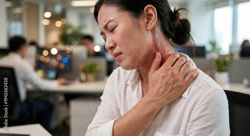 An Asian woman in an office setting closes her eyes in pain, gripping her inflamed neck and shoulder, clearly experiencing significant discomfort.