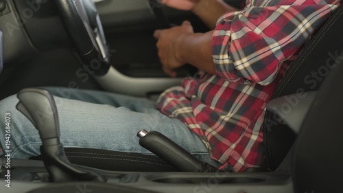 A man wearing a red plaid shirt is fastening his seatbelt in his private car. This is a safety precaution before driving.