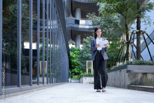 A professional businesswoman in a suit walks through the glass building office while looking at document embodying success and a modern work lifestyle outside an office building