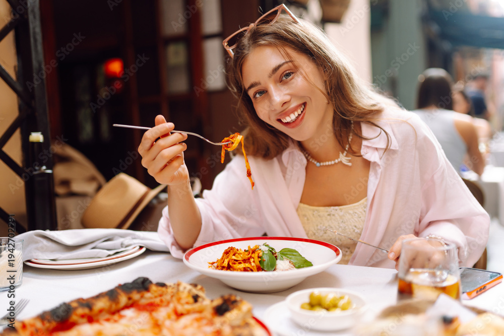 Obraz premium A happy woman eats fresh Italian pasta at a cafe table. A young tourist enjoys spaghetti on a sunny day on the terrace. Concept of food, enjoyment.