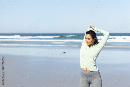 Asian woman stretching on beach with wet sand, wearing green top and grey leggings, copy space