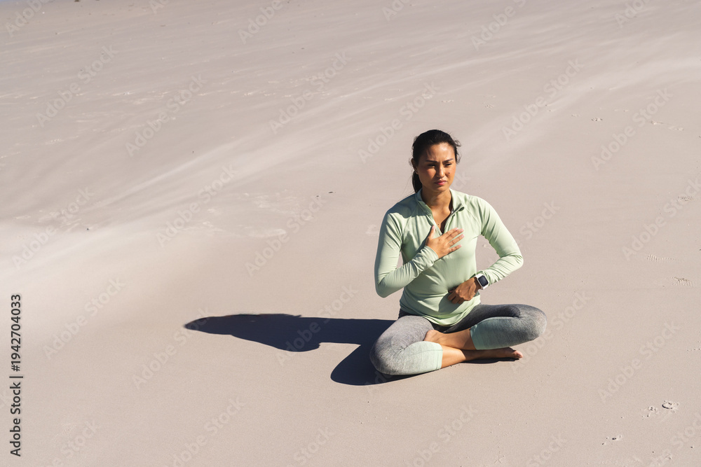 Fototapeta premium Mid-adult Asian woman sitting cross-legged on beach with smartwatch in green leggings, copy space