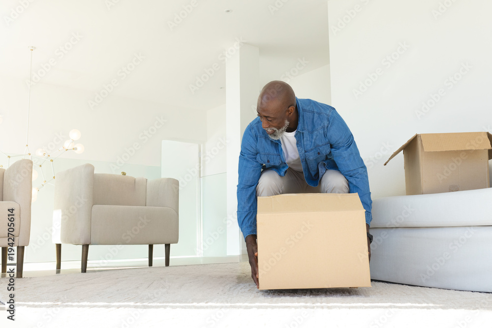Obraz premium Senior African American man handling moving box on rug in modern living room wearing denim jacket