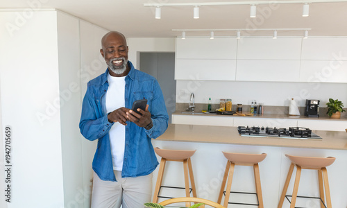 African American man smiling while holding smartphone in modern kitchen near island and stools