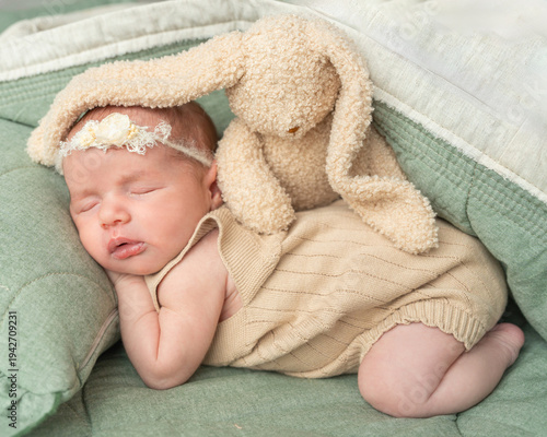 Sweet newborn baby girl sleeping peacefully with a plush bunny toy on soft green cushions. Tender studio portrait expressing innocence, childhood comfort and loving family atmosphere.