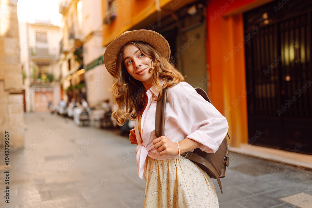 Obraz premium Beautiful tourist wearing hat and carrying backpack strolls through narrow streets of city on sunny day. Young woman enjoys the architecture and the city walk. A weekend, travel concept.