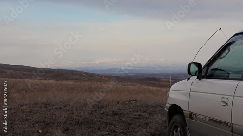 Early spring, mountain landscape, low clouds, a white car parked on a hill, looking at the horizon where snow-capped mountains are visible, the wind sways the dry grass