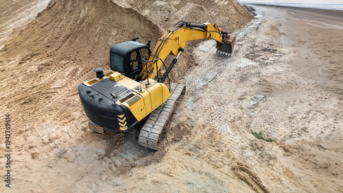 A large excavator is digging along the beach during the day. It is moving sand and shaping the area near the coastline
