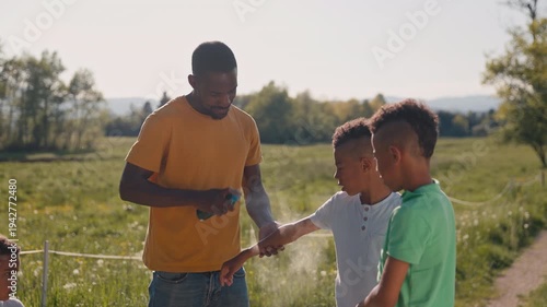 In the countryside, a father looking after his three sons, spraying the older twins with mosquito repellent before they play in nature, medium shot.