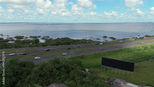 BR-290 Freeway interchange runs beside a lagoon with traffic, transmission towers and distant wind turbines in a wide aerial view, captured in D-Log with 10-bit color using a Hasselblad sensor. Brazil