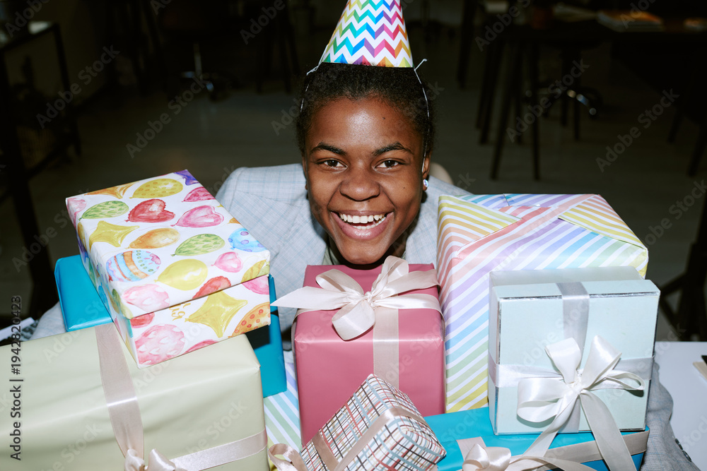 Obraz premium Portrait of Black girl wearing party hat smiling behind stack of wrapped birthday presents, showing joyful expression, sitting at table in indoor celebration setting