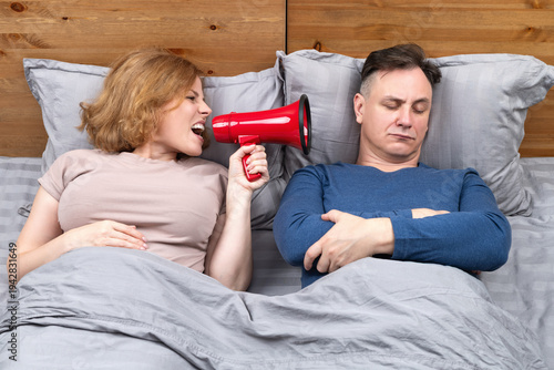 Bild auf Leinwand Funny wake up concept, woman shouting through red megaphone into sleeping man's ear in bed