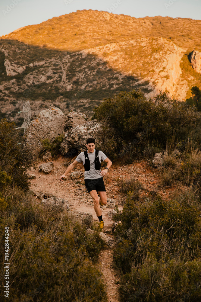 Obraz premium Young trail runner running along a steep mountain path surrounded by rugged scenery and the warm light of the sunset. Outdoor endurance training and adventure sport concept.