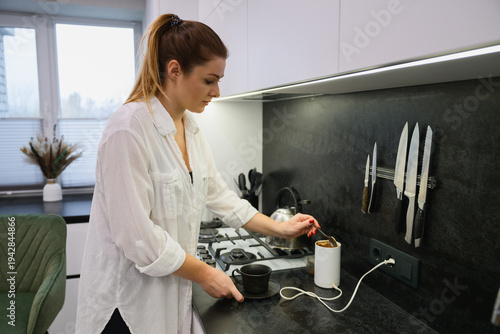 Young woman adding coffee beans to an electric grinder while preparing fresh coffee in a modern kitchen. Morning routine, homemade coffee preparation and everyday lifestyle.