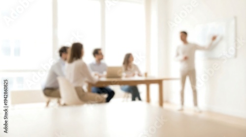 A blurred business meeting shows a man presenting to colleagues seated at a table in a brightly lit modern office setting.