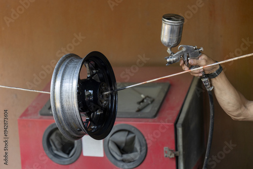 Focused professional painting black car wheel with spray gun. This automotive repair happens in garage workshop, applying new protective coating to metal rim