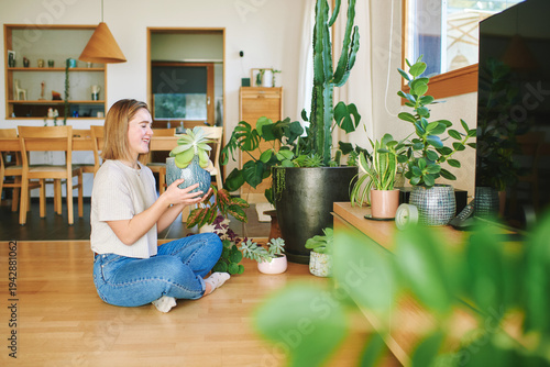 Young woman holding green house plant while sitting on floor in bright modern living room with many indoor plants cozy home lifestyle scene