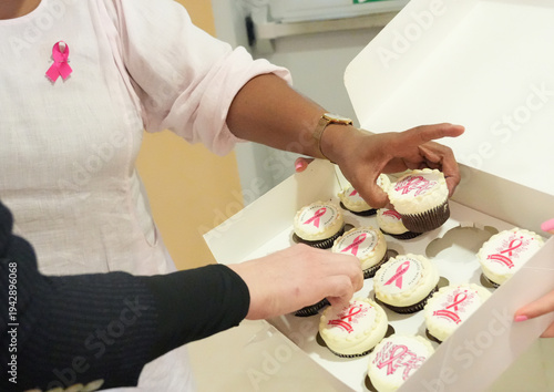 Participants from diverse backgrounds take cupcakes decorated with pink ribbon symbols during a breast cancer awareness event in a school hallway.