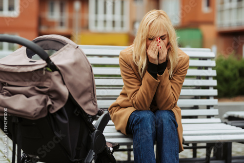 Upset young mother sitting on a park bench beside stroller covering her face in distress