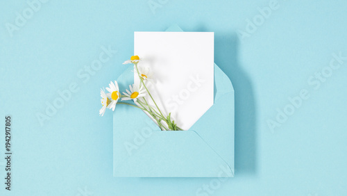 White chamomile flowers in a blue envelope with a blank greeting card on a pastel background. Minimalist spring flat lay, mockup for wedding invitation, Mother's Day, or romantic letter.