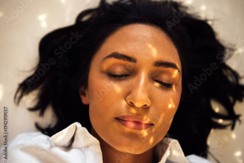 Asian woman with closed eyes, lying on soft surface, illuminated by gentle sunlight, creating a serene atmosphere, showcasing tranquility and peacefulness in a calm environment