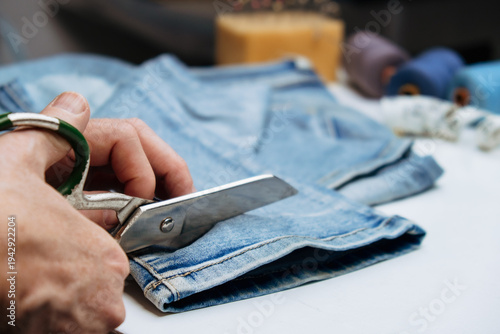 person is focused on cutting denim fabric with scissors on table. tailor cuts off extra length jeans with scissors. alteration, hemming, shortening or upgrade clothes. close up.