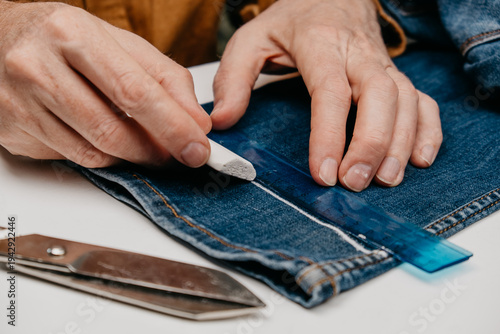 tailor marks denim fabric with chalk while using ruler for precision. scissors are placed nearby. this takes place in home workshop during day. closeup.