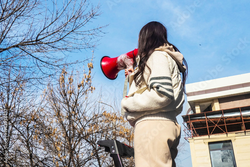 Young woman standing on a ladder and speaking through a megaphone during an outdoor street promotion. Public announcement in an urban environment with buildings and trees in the background.