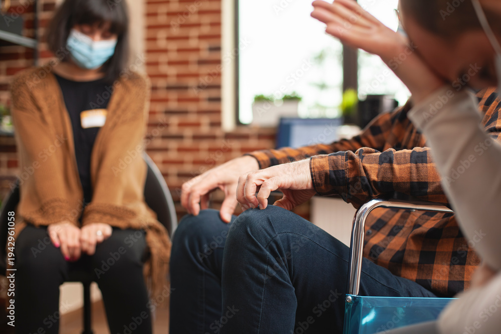 Fototapeta premium Closeup of male person touching his forehead, seated near elderly wheelchair user in brick wall room. Selective focus on hands of man in wheelchair as young woman in face mask sitting nearby.