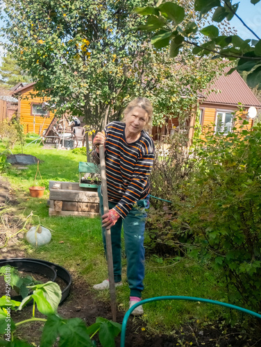 Mature woman gardener holding shovel in backyard. Senior person working in garden at countryside. Hobby, cultivation, agriculture and healthy lifestyle concept. Outdoor farming activity.