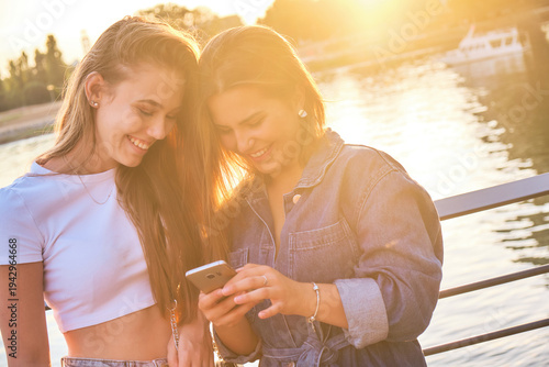 Two young women enjoying friendship moments and happy emotions while scrolling on a smartphone, sharing digital content outdoors during golden hour sunset by the river