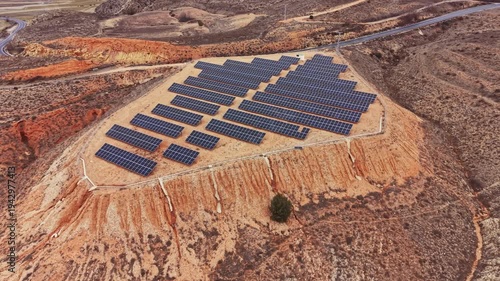 Solar panels cover a hillside in a remote location, arranged in rows under bright sunlight. The surrounding landscape shows dry earth and minimal vegetation.