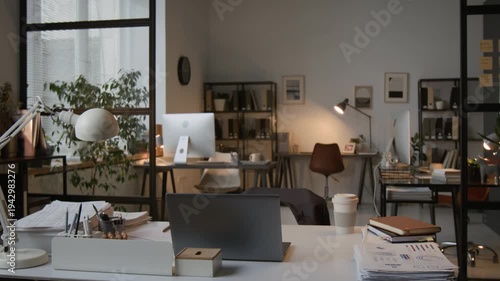 No people shot of modern office interior with several workstations, and white desk with laptop, documents, and lamp