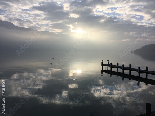 Peaceful view of Lake Annecy reflecting the surrounding mountains and cloudy sky. The calm water mirrors the alpine peaks and dramatic clouds, creating a serene and atmospheric landscape. 