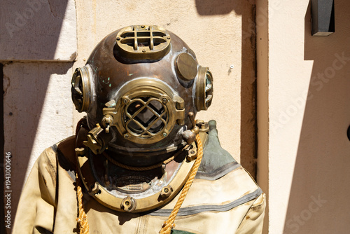Antique Brass Diving Helmet On Vintage Diving Suit Displayed Against Sunlit Wall Maritime Relic