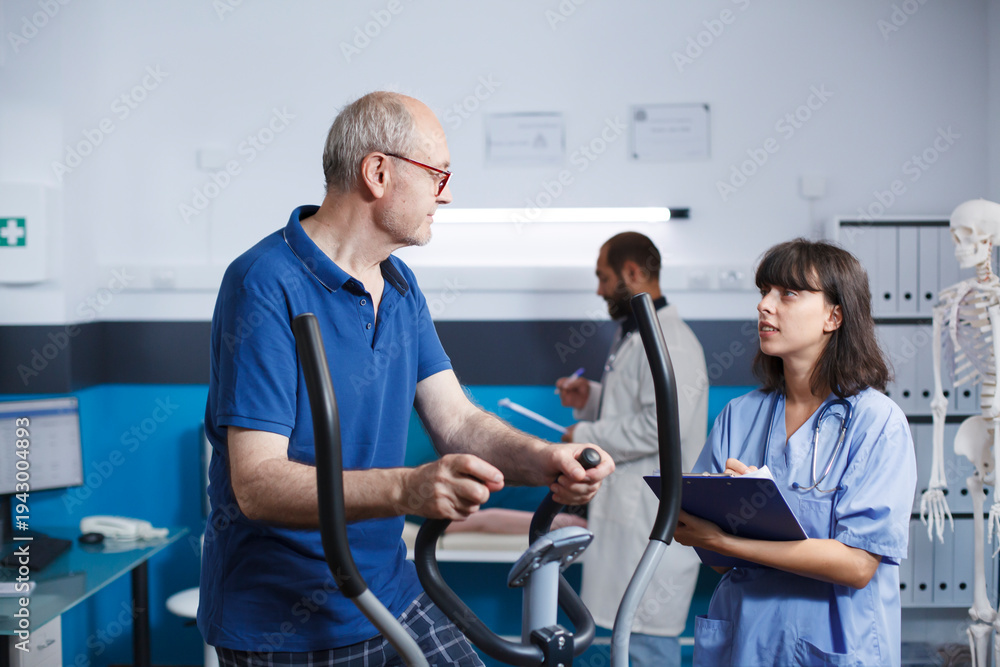 Fototapeta premium Elderly patient is seen receiving physiotherapy on stationary bike, assisted by female nurse dressed in blue scrubs. Senior man is getting rehabilitative care with help from health professional.