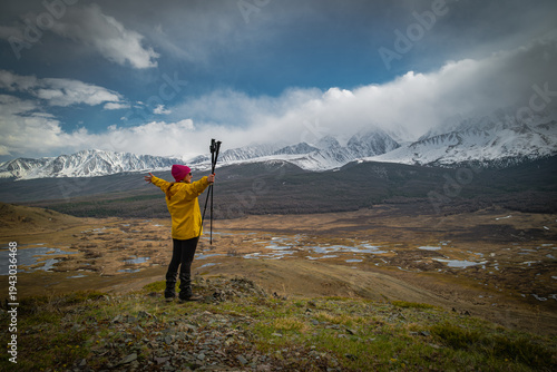 Back view of happy female hiker in yellow jacket with trekking poles raising arms in success standing on mountain edge overlooking scenic highland valley with snow peaks for freedom.